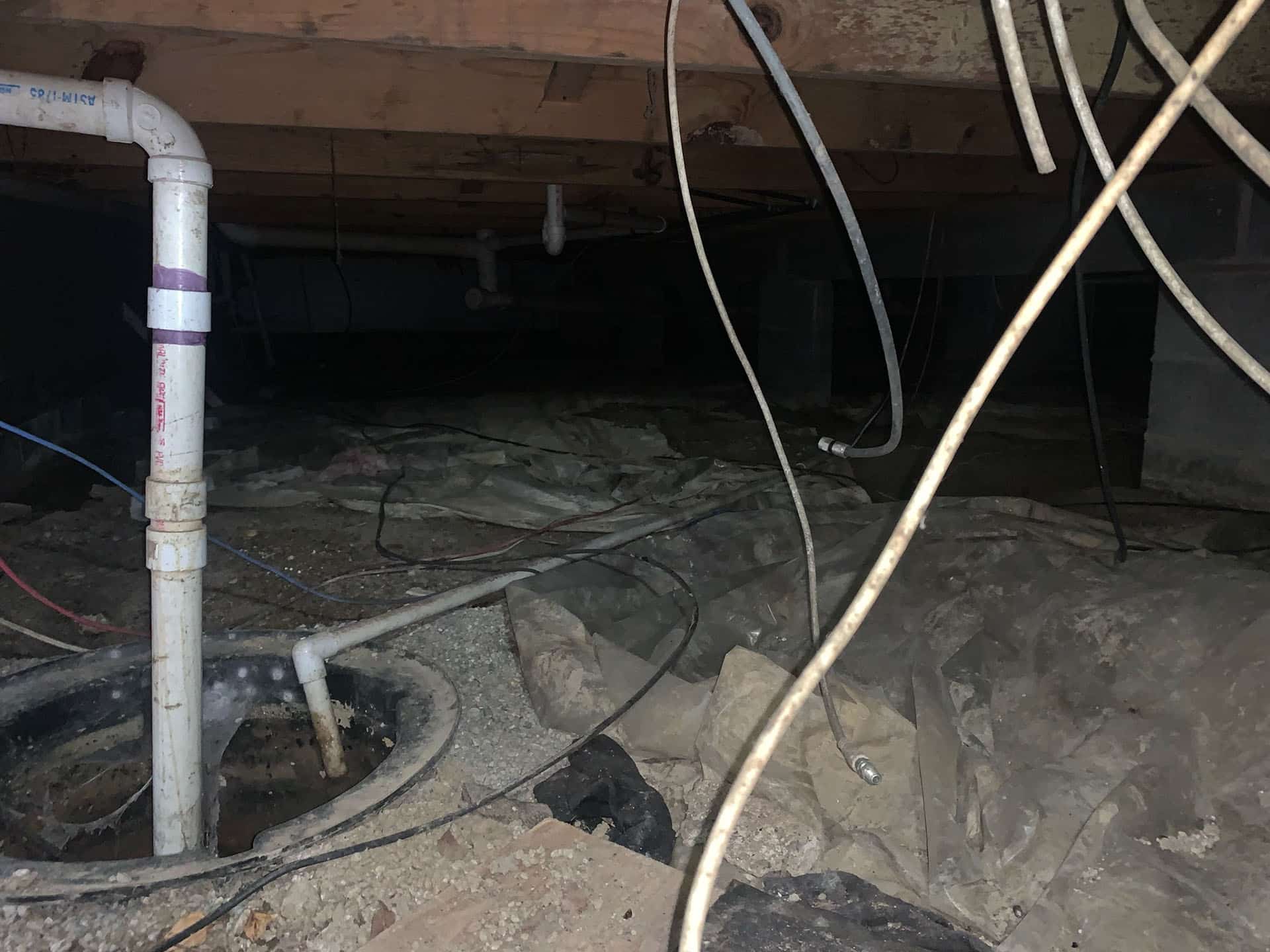 A dimly lit crawl space under a building with exposed pipes, concrete blocks, dirt floor, and plastic sheeting covering parts of the ground.