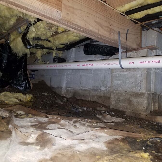 Crawl space with exposed insulation, ductwork, and a white PVC pipe running along a concrete block wall, with dirt and debris on the ground.