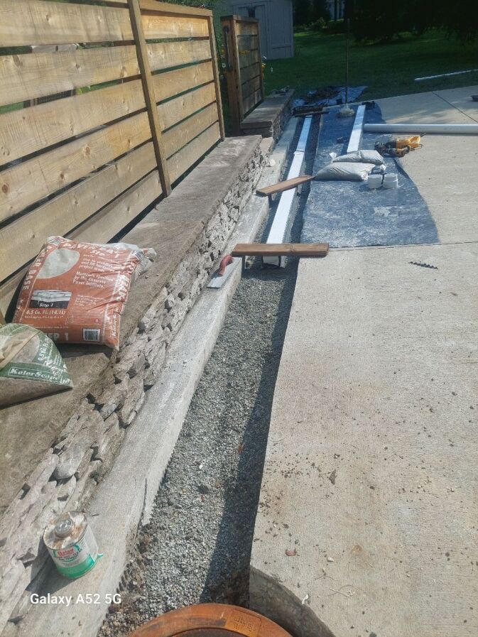 A partially constructed stone wall next to a wooden fence with bags of construction materials, tools, and gravel along the base.