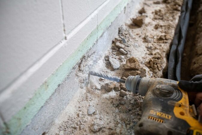 A person uses a DeWalt rotary hammer drill to bore into the base of an interior concrete wall, creating dust and debris along the foundation.