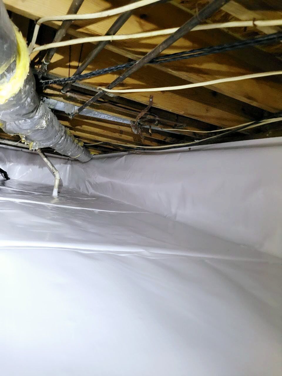 Exposed pipes and wires run along the wooden ceiling of a crawl space lined with white plastic sheeting.