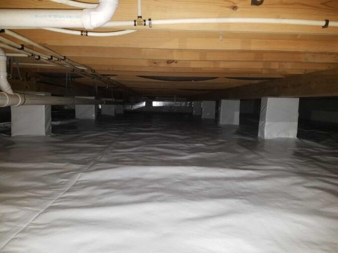 A clean, encapsulated crawl space with a plastic vapor barrier covering the floor and piers, visible plumbing pipes, and wooden floor joists above.