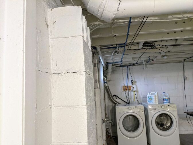 A basement with visible cracks in a cement wall, exposed pipes and wiring on the ceiling, and two front-loading laundry machines next to detergent containers.