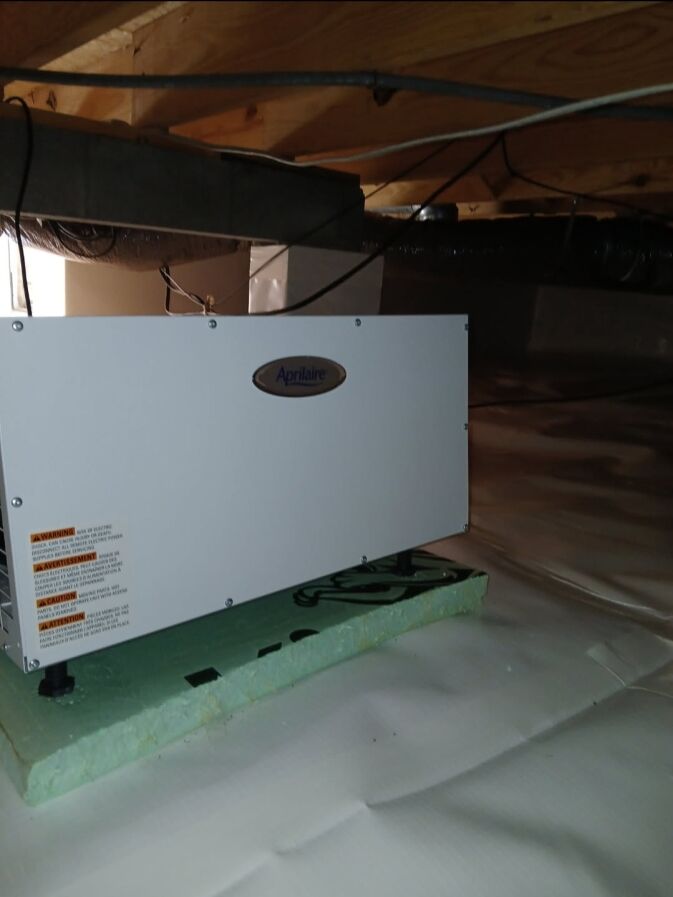 A white Aprilaire dehumidifier is installed on a foam platform in a clean, sealed crawl space with plastic vapor barrier and exposed wooden beams above.