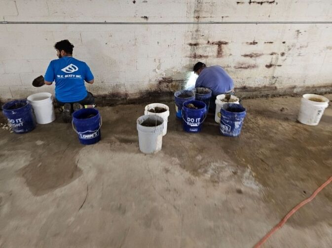 Two workers are repairing a wall, surrounded by several blue and white buckets filled with concrete or mortar material on a concrete floor.