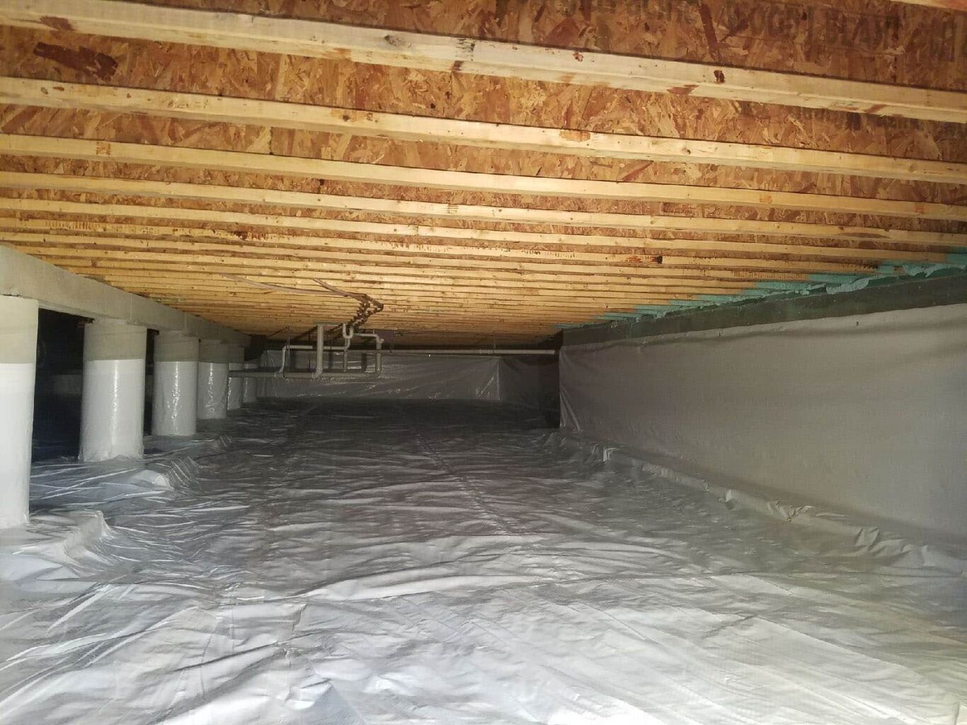 A crawl space with a plastic vapor barrier covering the floor, exposed wooden joists overhead, and foundation pillars along the sides.