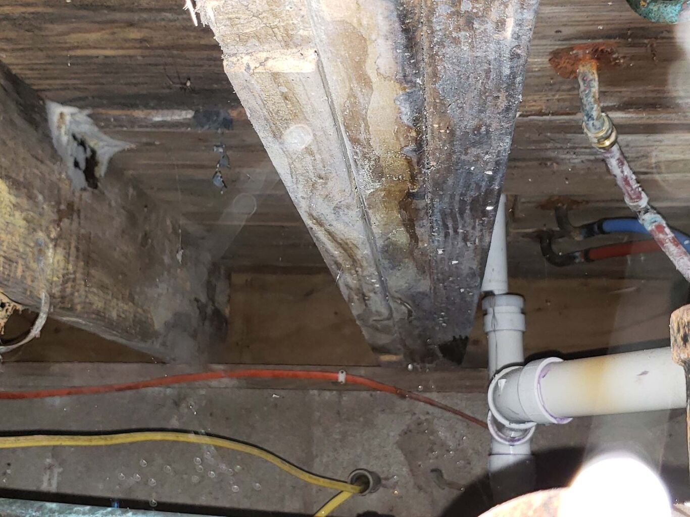 Exposed wooden beam with signs of water damage and mold next to plumbing pipes and electrical wires in a basement or crawl space.