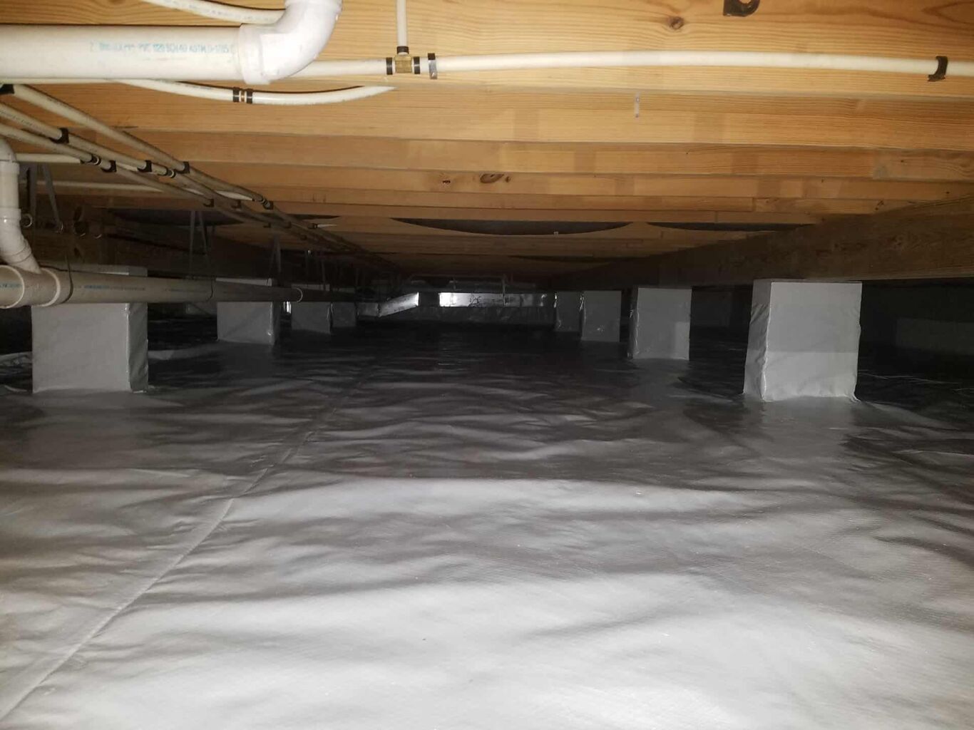 A clean, encapsulated crawl space with a plastic vapor barrier covering the floor and piers, visible plumbing pipes, and wooden floor joists above.