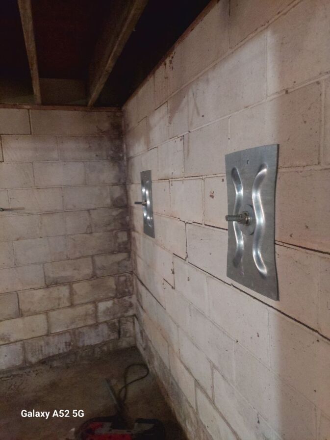 Two steel wall anchors installed on a basement concrete block wall, with visible bolts securing them in place; exposed ceiling joists are overhead.