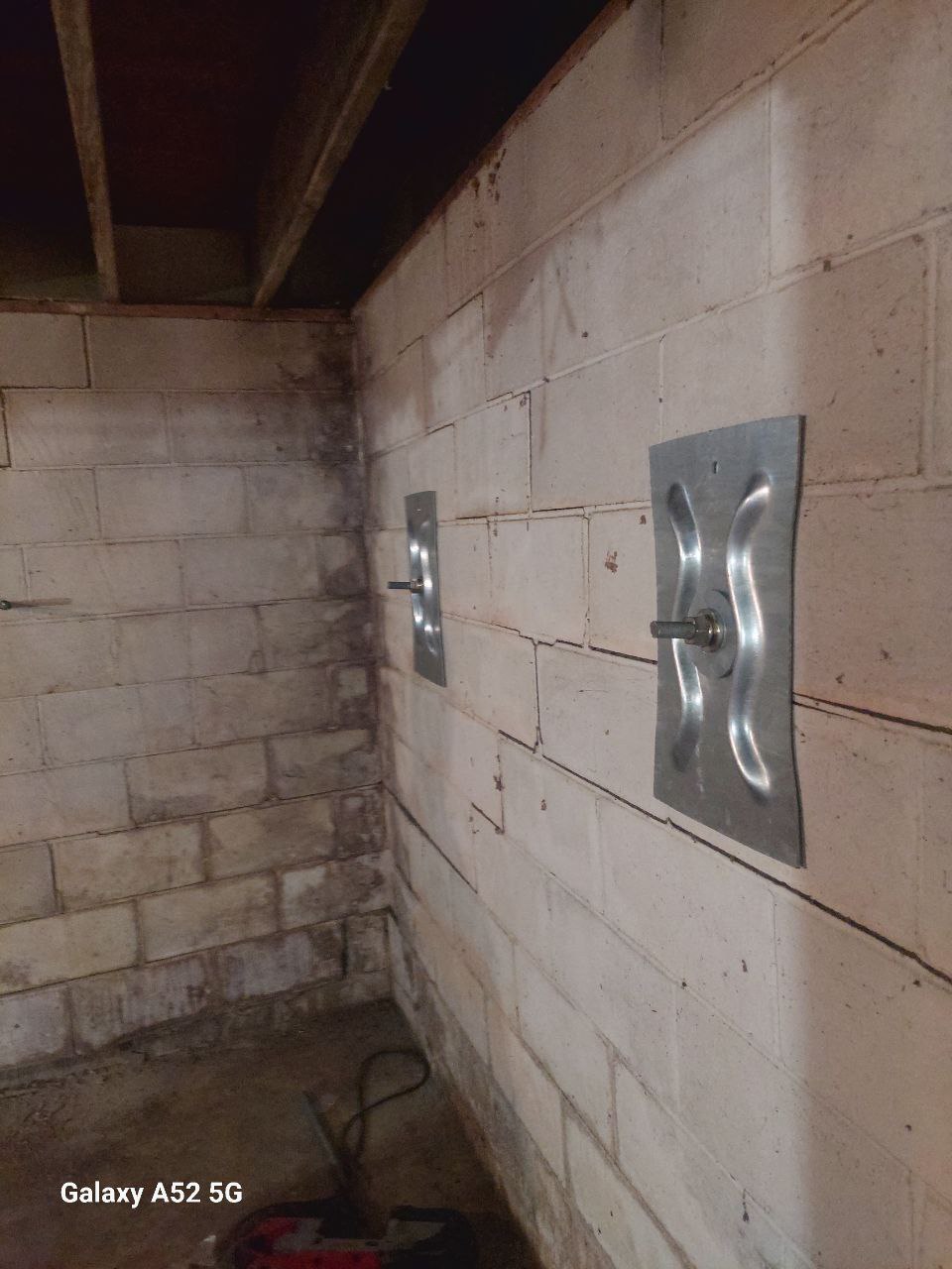 Two steel wall anchors installed on a basement concrete block wall, with visible bolts securing them in place; exposed ceiling joists are overhead.