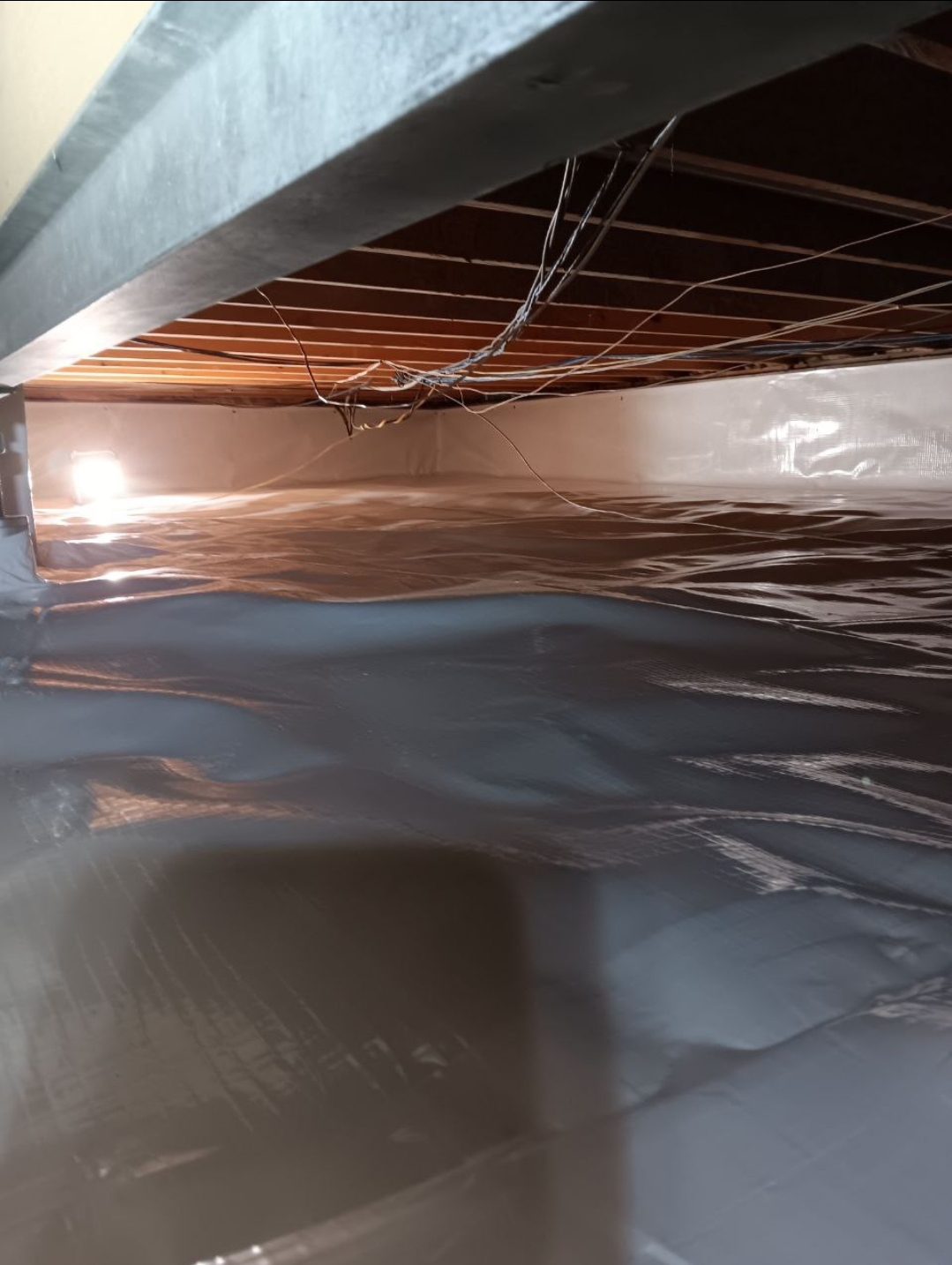 A crawl space with a plastic vapor barrier covering the floor and lower walls, exposed wooden beams, and some hanging wiring visible overhead.