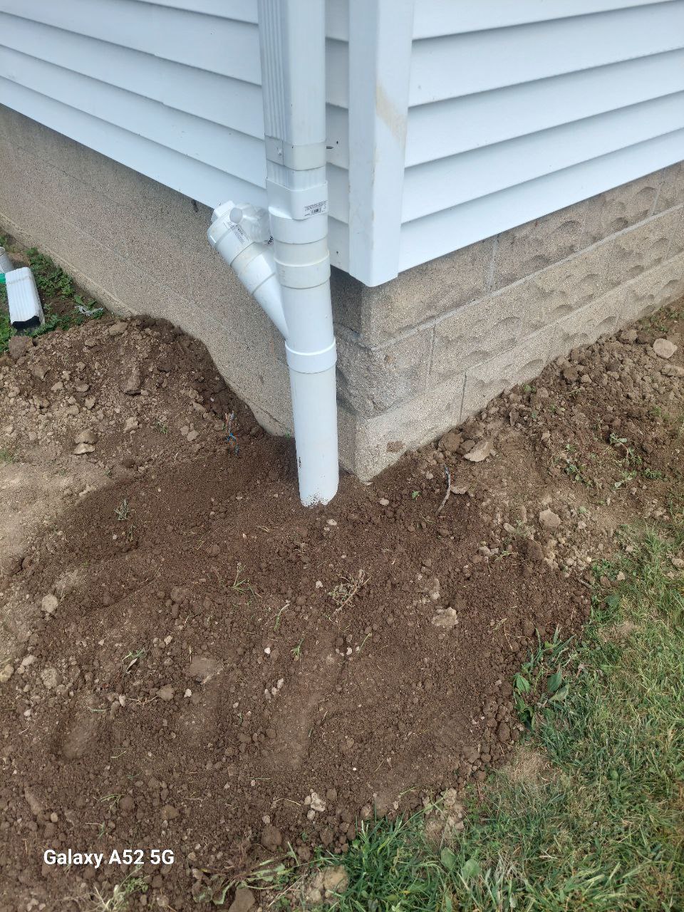 A section of house siding with a downspout drains into the ground; soil appears freshly disturbed around the foundation.