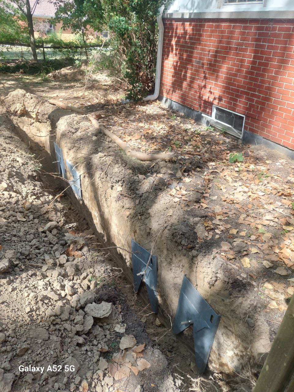 A deep trench with metal plates along one side runs parallel to a brick house, with soil and leaves scattered around the construction site.