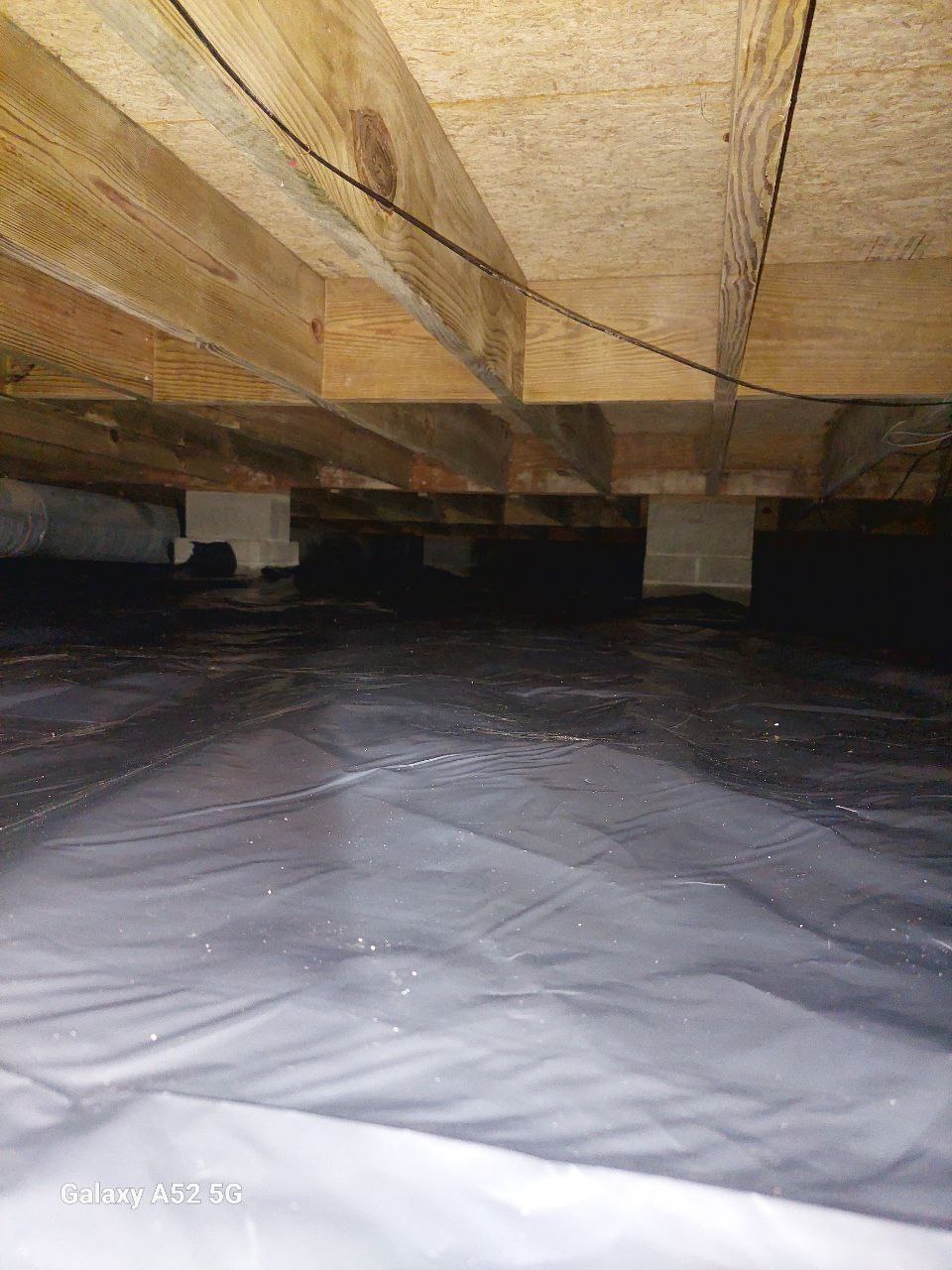 View of a crawl space under a building, showing wooden floor joists above and a plastic vapor barrier covering the ground.