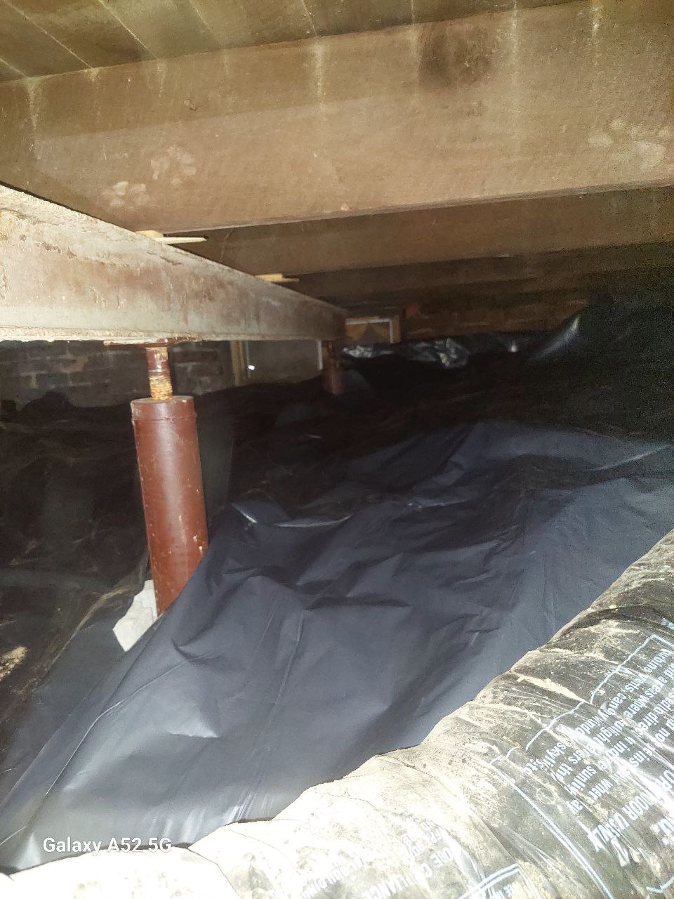 A crawl space with exposed wooden beams, metal supports, plastic vapor barrier on the ground, and some insulation visible in the foreground.