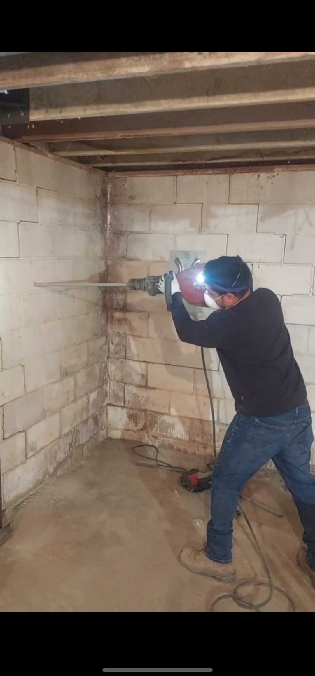 Person wearing safety gear uses a power tool to cut into a cinder block basement wall, with debris on the floor and exposed ceiling joists above.
