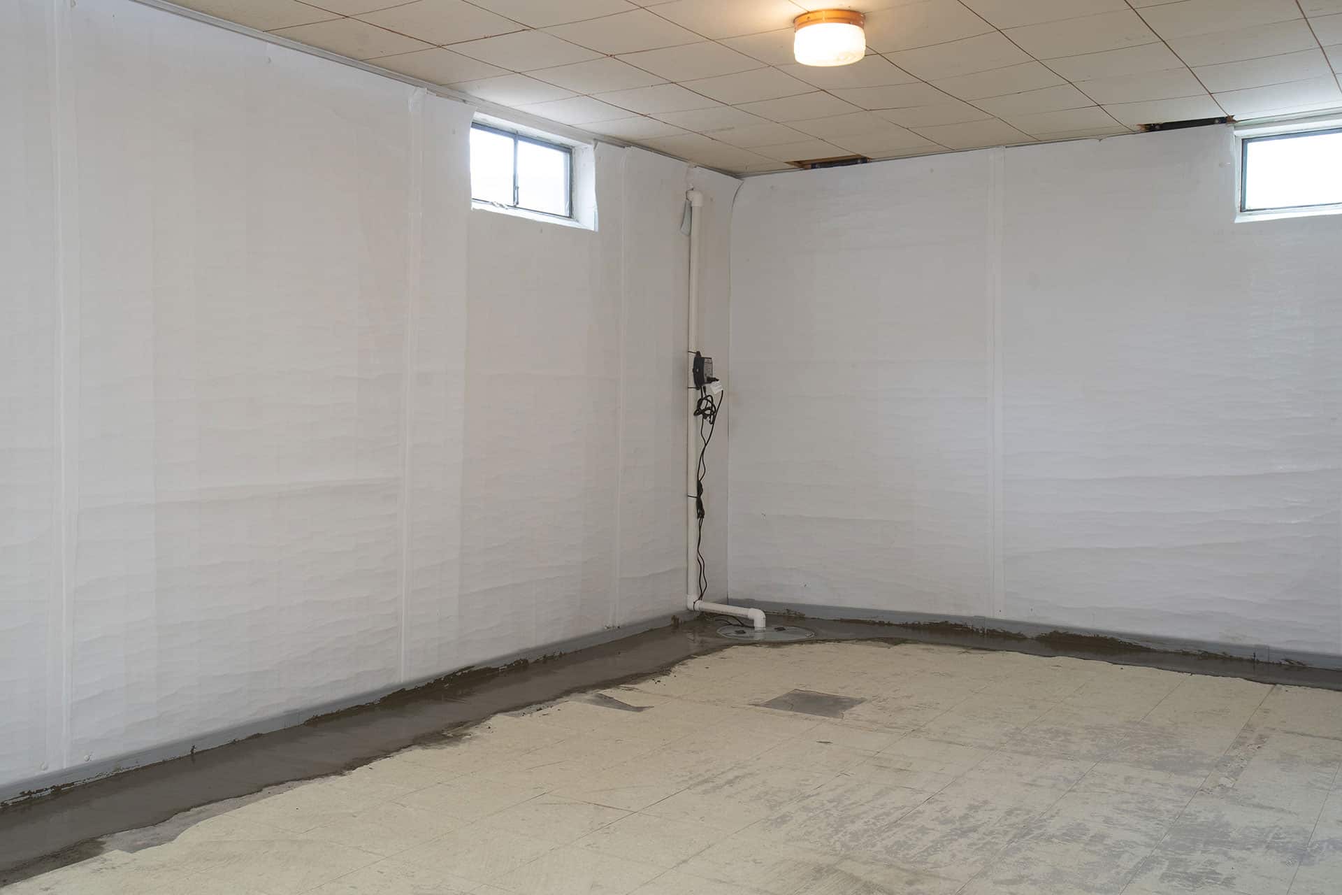 An empty basement room with white walls, small windows near the ceiling, exposed ceiling tiles, and unfinished flooring. Electrical equipment is mounted in the corner.