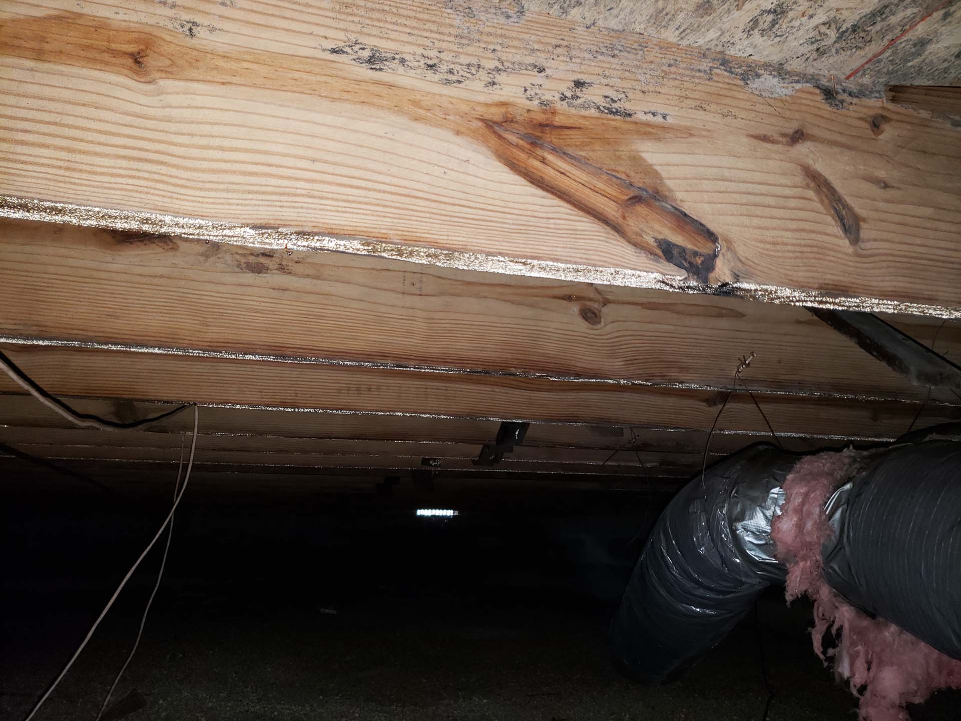 Crawl space with wooden beams, exposed insulation, metal ductwork, and some signs of moisture damage or mold on the wood.