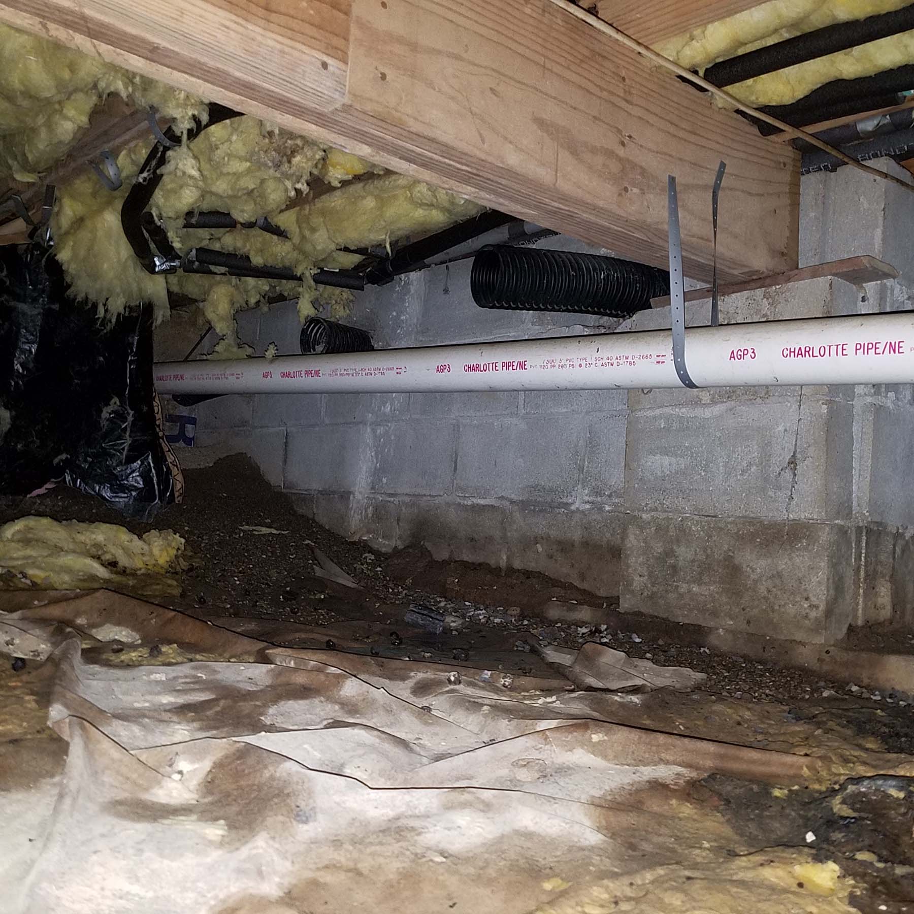 Crawl space with exposed insulation, ductwork, and a white PVC pipe running along a concrete block wall, with dirt and debris on the ground.