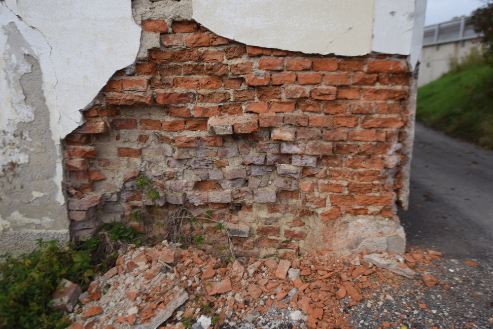 A deteriorating brick wall with significant plaster loss, revealing exposed red bricks and loose debris on the ground.