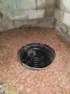 A black sump pump basin is installed in a basement corner, surrounded by gravel and unfinished concrete block walls.