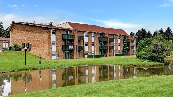 Three-story brick apartment building with balconies is reflected in a small pond, surrounded by green grass and trees under a blue sky.