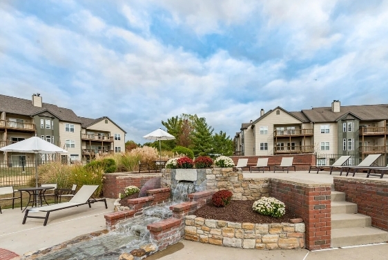 Apartment complex courtyard featuring a small waterfall, landscaped flower beds, lounge chairs, tables with umbrellas, and multi-story residential buildings in the background.